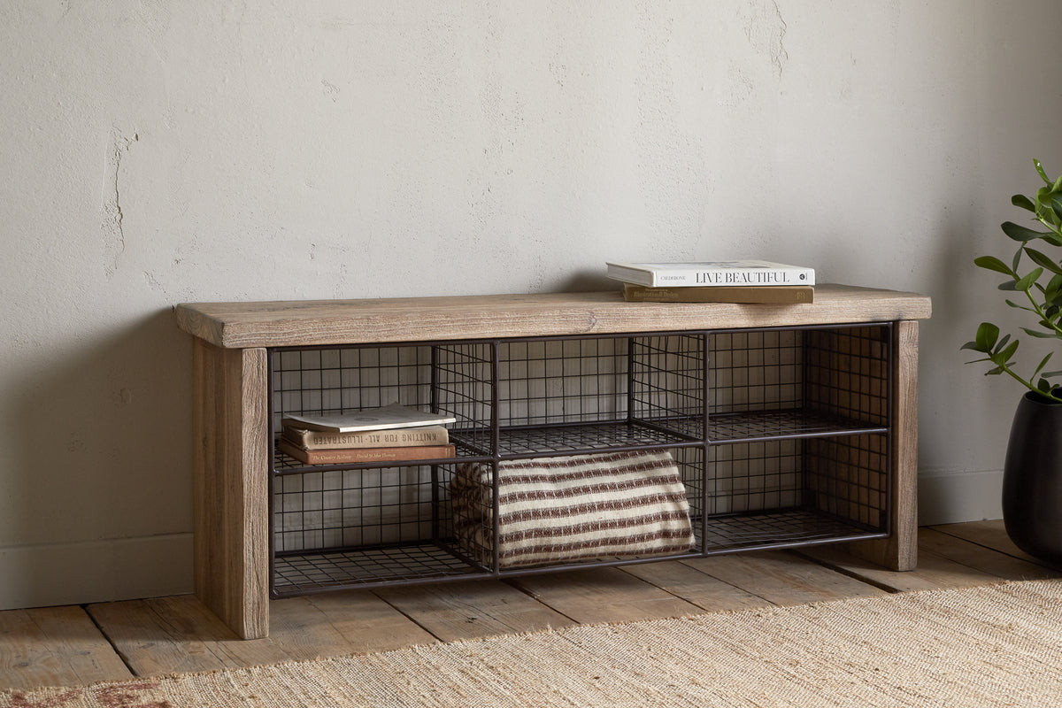 The nkuku Abha Reclaimed Wood & Iron Storage Bench - Natural holds a folded striped blanket and books, set against a pale wall by a potted plant on a woven rug—ideal as an entryway organizer.