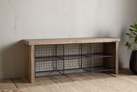 The nkuku Abha Reclaimed Wood & Iron Storage Bench – Natural is a versatile entryway organizer featuring six black wire baskets for shoe storage, set on a wooden floor beside a potted green plant.