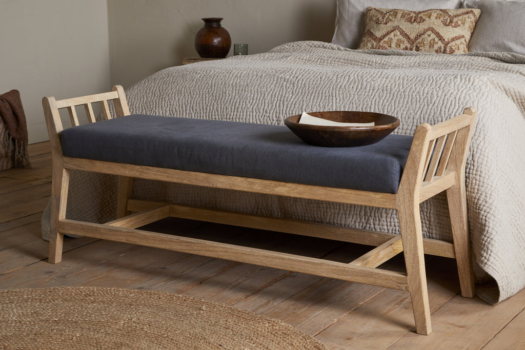 The Avanthi Upholstered Sleigh Bench by nkuku, featuring a charcoal cushion and carved wooden frame, sits at the foot of a neatly made bed with textured bedding in a cozy, neutral-toned bedroom.