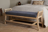 The Avanthi Upholstered Sleigh Bench by nkuku, featuring a charcoal cushioned seat and carved wooden frame, sits at the foot of a neatly made bed in a cozy bedroom with neutral tones and rustic decor.