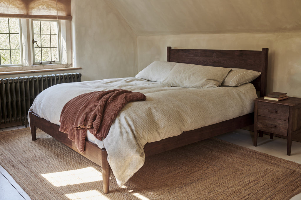 A cozy bedroom features the nkuku Anbu Acacia Bed in Washed Walnut, beige bedding, and a rust blanket at the corner. A wooden nightstand with books sits beside the bed as sunlight filters through window blinds.