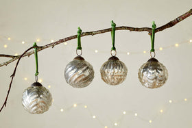 The Adoya Baubles - Aged Nickel (Set of 4) by nkuku hang from a branch with a green velvet tie, surrounded by warm white fairy lights and a soft backdrop. Their ribbed texture adds cozy charm to your festive décor.