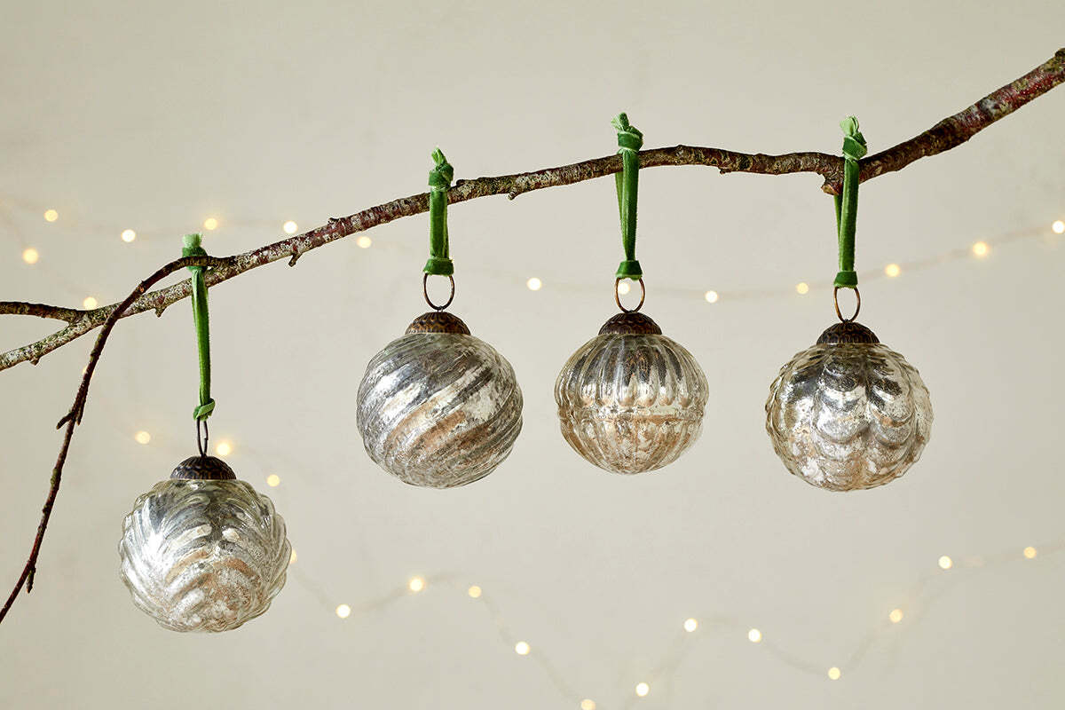 The Adoya Baubles - Aged Nickel (Set of 4) by nkuku hang from a branch with a green velvet tie, surrounded by warm white fairy lights and a soft backdrop. Their ribbed texture adds cozy charm to your festive décor.