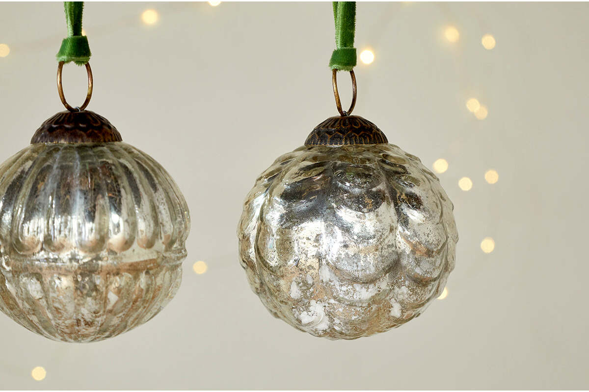 The Adoya Baubles - Aged Nickel (Set of 4) by nkuku hang from green velvet ties, with textured patterns. Warm fairy lights glow softly in the blurred background, adding a cozy and festive touch to your décor.