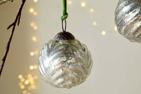 The Adoya Baubles - Aged Nickel (Set of 4) by nkuku, featuring vintage-style ribbed glass with a leafy pattern and a green velvet tie, hang amid festive decor with soft yellow fairy lights for a cozy holiday ambiance.