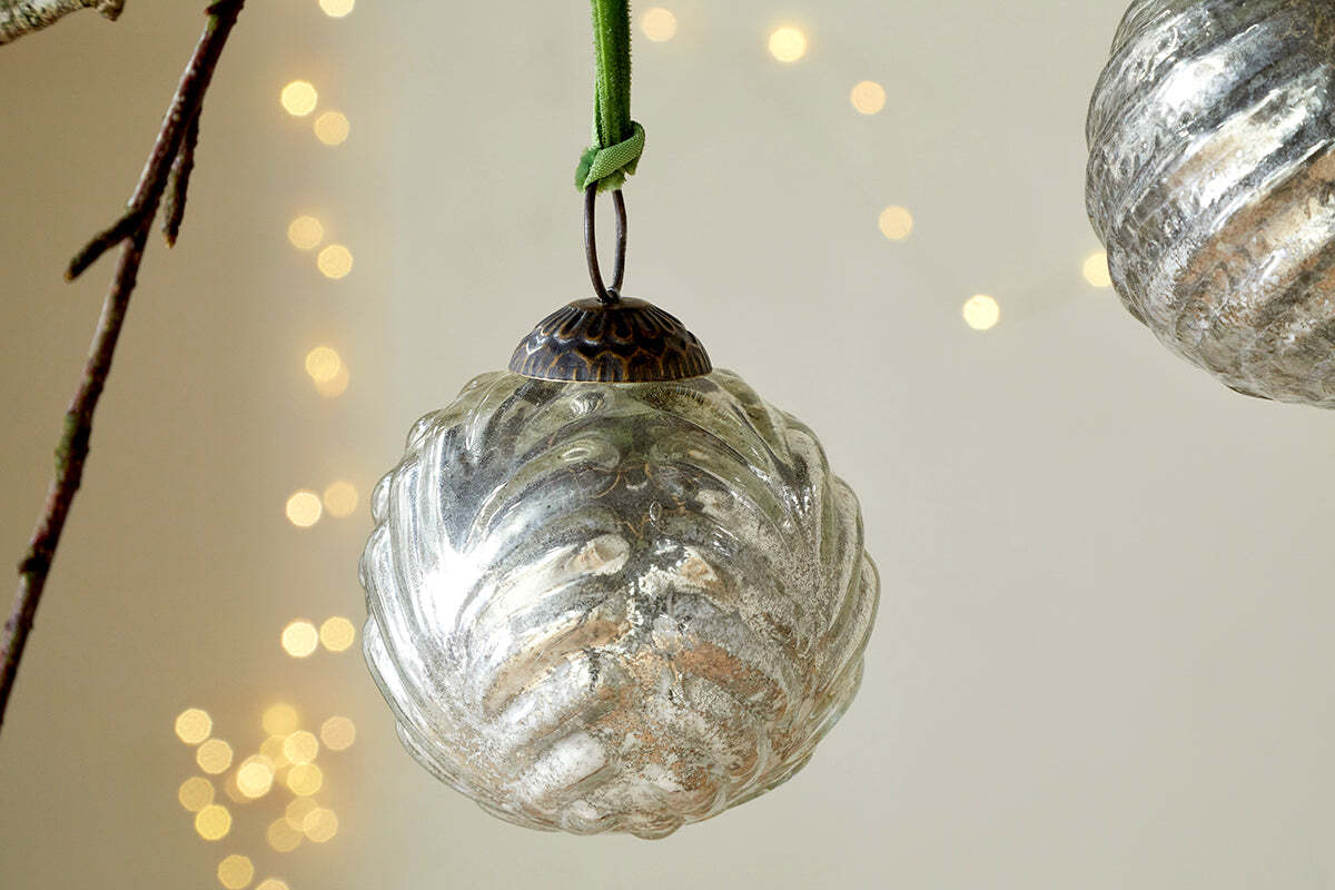 The Adoya Baubles - Aged Nickel (Set of 4) by nkuku, featuring vintage-style ribbed glass with a leafy pattern and a green velvet tie, hang amid festive decor with soft yellow fairy lights for a cozy holiday ambiance.
