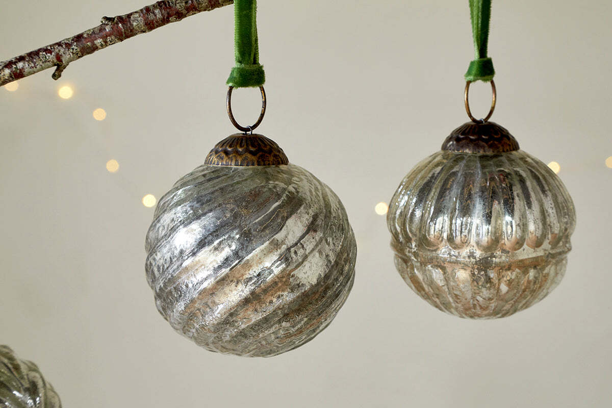 Four vintage-inspired Adoya Baubles in aged nickel from nkuku hang by green velvet ties. Grooved textures and soft fairy lights in the background add a cozy, festive touch to your holiday decor.