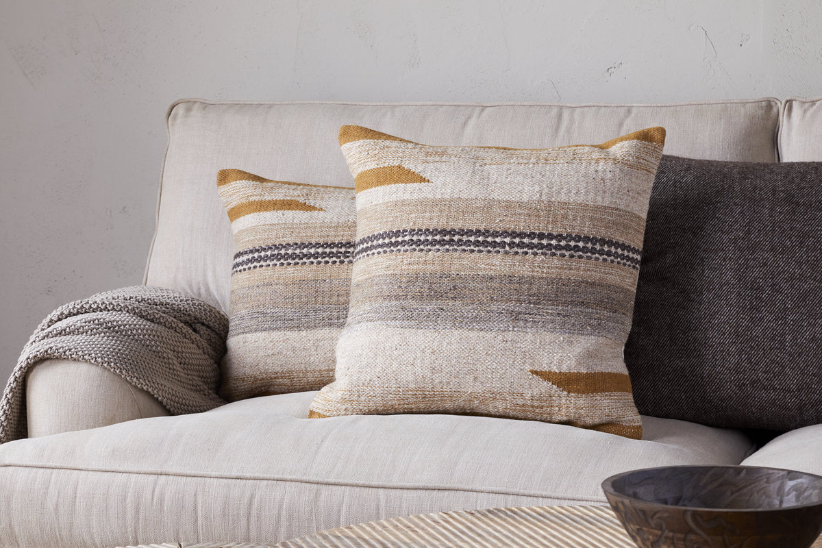 A beige sofa features two nkuku Aravali Wool Cushion Covers in ochre and earthy tones, a gray textured blanket over the arm, a dark gray pillow behind, and a wooden bowl on a light-colored table in front.