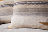 Close-up of two nkuku Aravali Wool Cushion Covers in ochre, featuring a woven design with horizontal beige, brown, and gray stripes. The covers have a rustic wool texture and rest on a light fabric surface.