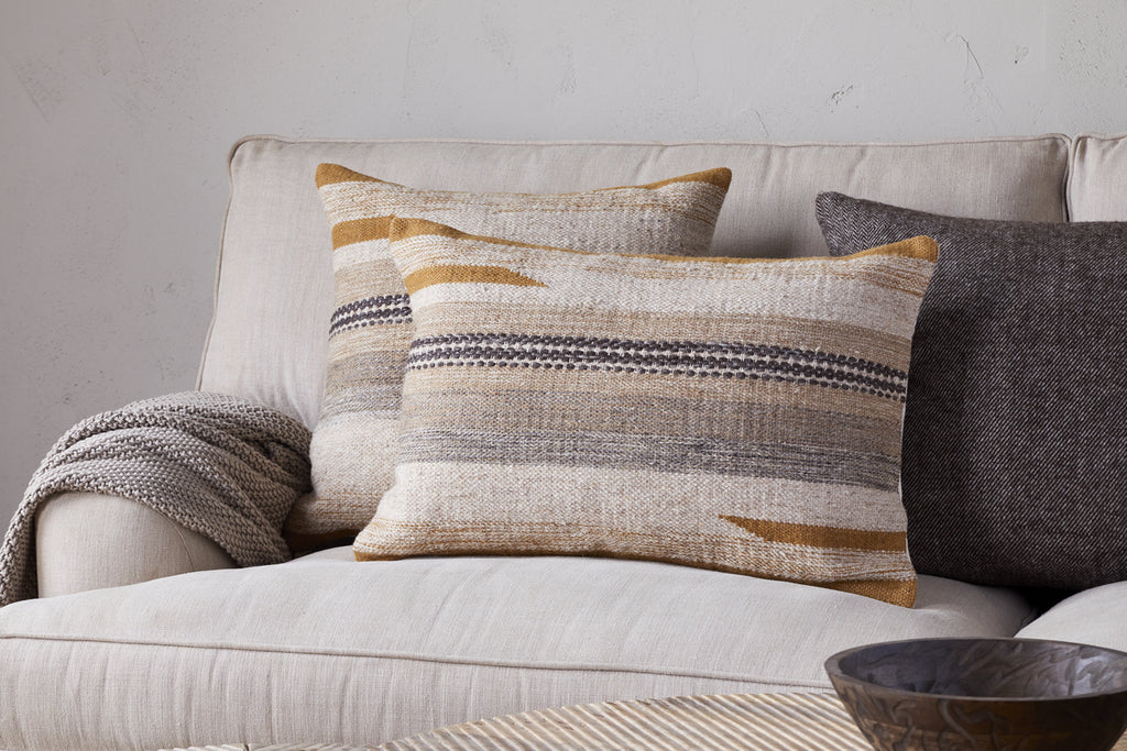 Two patterned beige pillows with brown and gray stripes rest on a light sofa. A gray knitted throw is draped over the armrest, and the nkuku Aravali Wool Cushion Cover in Ochre sits in the corner. A wooden bowl is visible in the foreground.