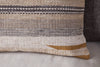 The nkuku Aravali Wool Cushion Cover - Ochre features beige, brown, and gray horizontal stripes with textured weaving, shown in a close-up resting against a gray pillow on a light surface.