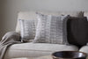 A beige sofa features two patterned gray-and-white cushion covers, an nkuku Aahna Wool Cushion Cover in charcoal, and a textured knit blanket. In the foreground, a dark wooden bowl rests on a light-colored surface.