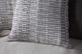 A close-up shows the nkuku Aahna Wool Cushion Cover - Charcoal, featuring a woven gray and white geometric pattern on a light sofa, with another dark pillow behind it. The cover is crafted from wool and recycled polyester.