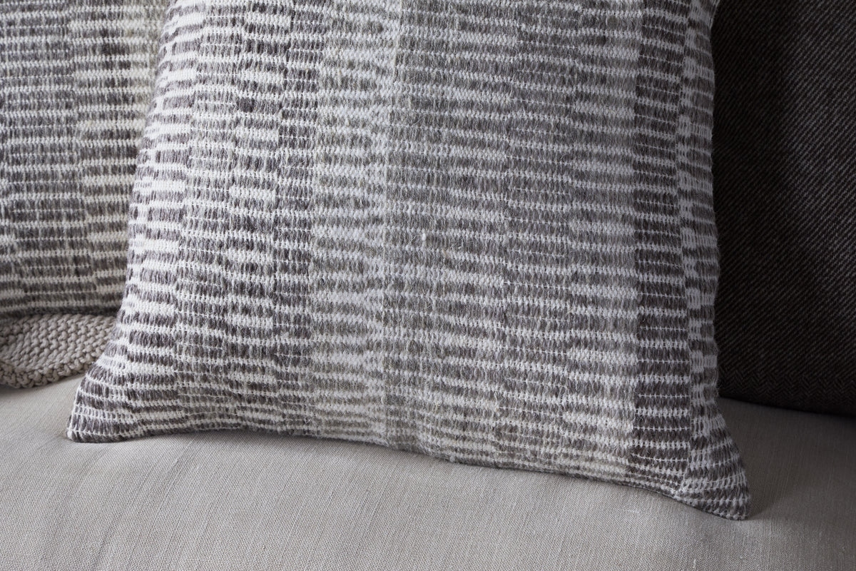 A close-up shows the nkuku Aahna Wool Cushion Cover - Charcoal, featuring a woven gray and white geometric pattern on a light sofa, with another dark pillow behind it. The cover is crafted from wool and recycled polyester.