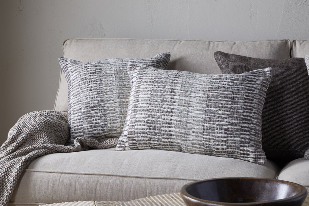 A beige sofa features two nkuku Aahna Wool Cushion Covers in charcoal, a textured knit blanket draped over one arm, and a dark brown pillow in the corner. A wooden bowl sits on a table in the foreground.
