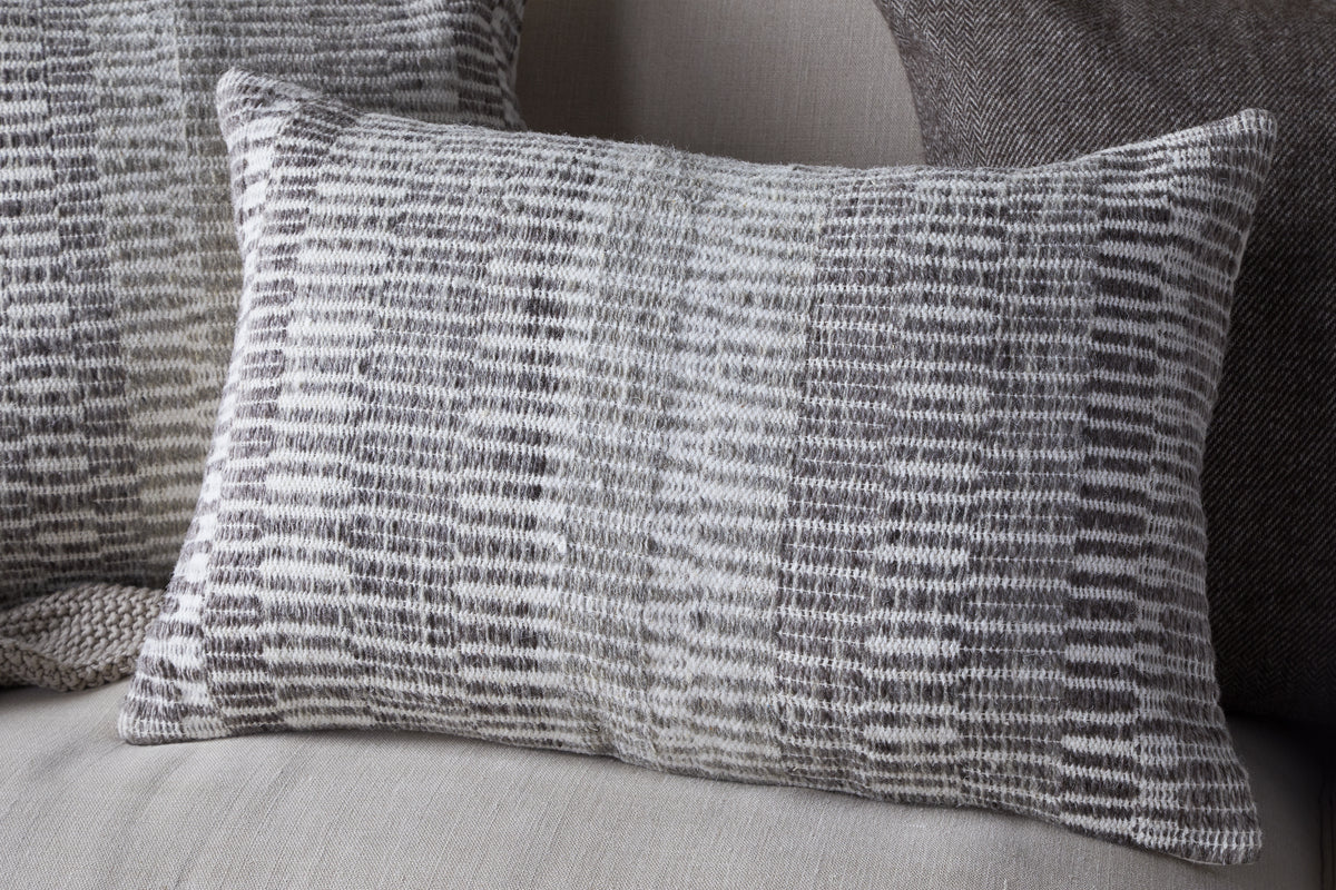 The nkuku Aahna Wool Cushion Cover in Charcoal features a textured, woven design in gray and white wool and recycled polyester, styled here on a light fabric sofa with coordinating cushions in the background.