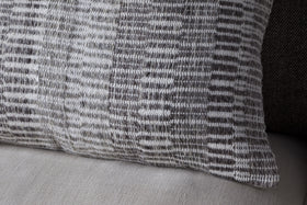 A close-up of the nkuku Aahna Wool Cushion Cover in charcoal, featuring a textured woven design in gray and white wool and recycled polyester, displayed on a light beige fabric surface.