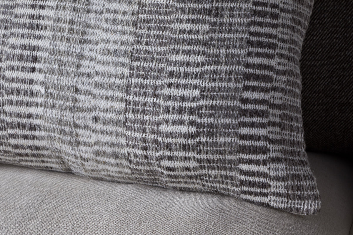 A close-up of the nkuku Aahna Wool Cushion Cover in charcoal, featuring a textured woven design in gray and white wool and recycled polyester, displayed on a light beige fabric surface.
