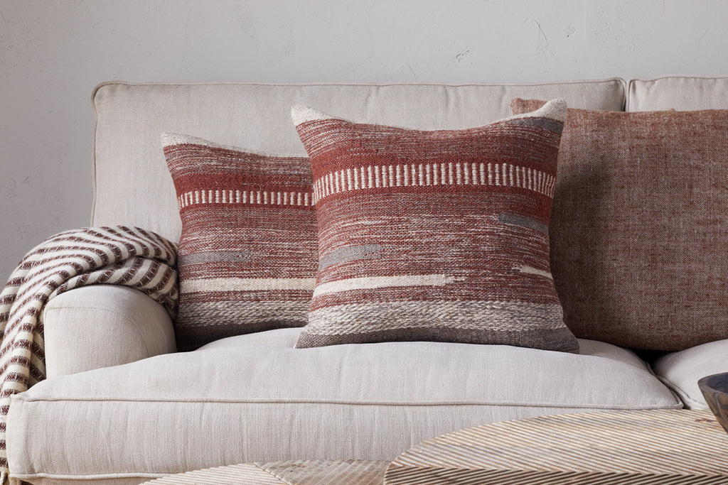 A beige sofa with two red and beige geometric pillows, an nkuku Aakash Wool Cushion Cover in rust, and a striped knit throw blanket draped over the armrest. A light-colored coffee table sits in the foreground.