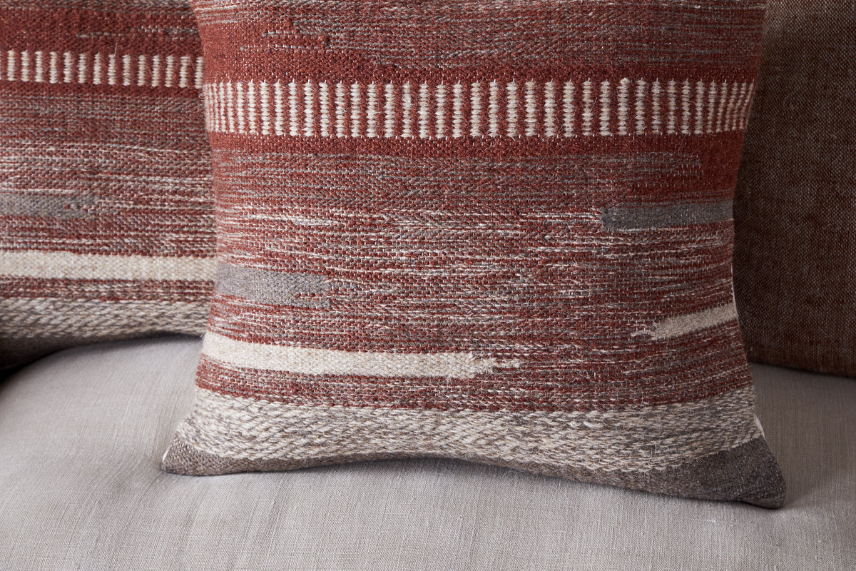 The Aakash Wool Cushion Cover - Rust by nkuku features earthy tones of rust, beige, and gray in textured, woven horizontal patterns, shown here on a light gray upholstered surface.