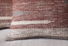 Close-up of the nkuku Aakash Wool Cushion Cover in rust, featuring textured horizontal stripes in earthy brown, beige, and cream tones, shown on a light gray fabric surface.