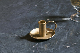 The Amri Iron Candlestick by nkuku, finished in antique brass, sits on a matching round dish atop dark veined marble. To the right, the base of a partially visible goblet or glass casts a soft shadow.