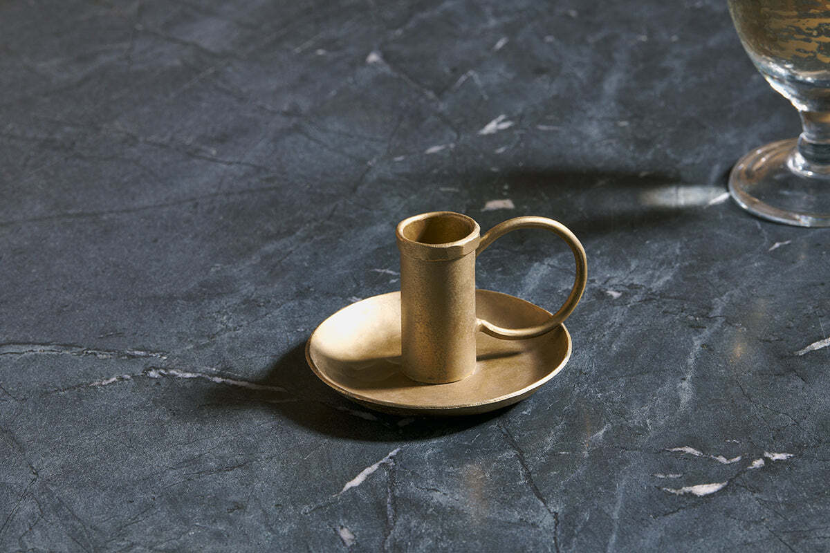 The Amri Iron Candlestick by nkuku, finished in antique brass, sits on a matching round dish atop dark veined marble. To the right, the base of a partially visible goblet or glass casts a soft shadow.