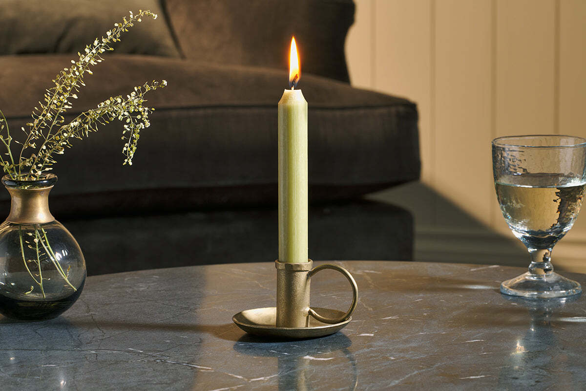 An Amri Iron Candlestick in Antique Brass by nkuku sits lit on a gray marble table, beside a small glass vase with sprigs and a half-filled water glass. A dark sofa and beige paneled wall create a cozy, warm atmosphere.
