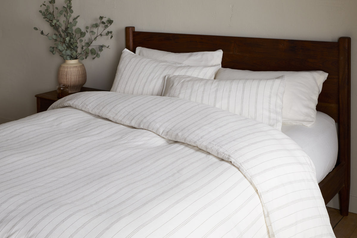 A neatly made bed with white and light gray striped bedding, four pillows, and a wooden headboard features the nkuku Anta Hemp & Cotton Duvet Cover in Sage Green. A ceramic vase with green branches sits on the bedside table.