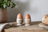 Two brown eggs in nkukus Awa Wooden Egg Cup - Natural (Set of 2) sit on a rustic breakfast table with tableware, bread, a potted plant, and a cloth against a light textured wall.