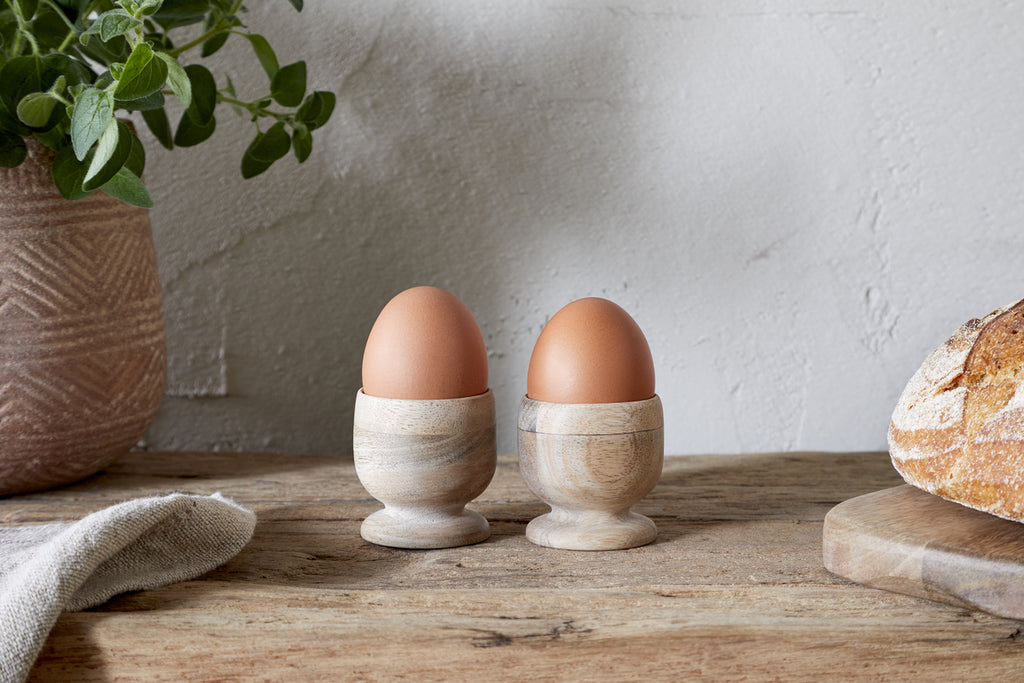 Two brown eggs in nkukus Awa Wooden Egg Cup - Natural (Set of 2) sit on a rustic breakfast table with tableware, bread, a potted plant, and a cloth against a light textured wall.