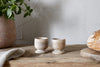 The nkuku Awa Wooden Egg Cups (Set of 2, Natural) sit on a rustic breakfast table beside a loaf of bread, with a potted plant and folded cloth completing the inviting scene.