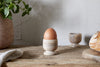 A brown egg sits upright in the nkuku Awa Wooden Egg Cup - Natural (Set of 2) on a rustic table, surrounded by another empty cup, bread, a leafy potted plant, and a cloth for charming morning tableware.