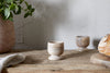 The Awa Wooden Egg Cup - Natural (Set of 2) by nkuku sits on a rustic breakfast table beside a round loaf, potted plant, and folded cloth, against a textured light gray wall.