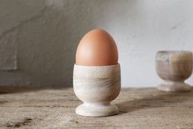 A brown egg stands in the nkuku Awa Wooden Egg Cup - Natural (Set of 2), part of stylish tableware, on a rustic wooden surface with the second cup softly blurred in the background.