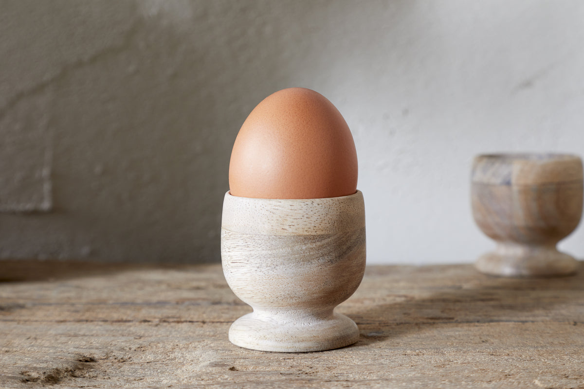 A brown egg stands in the nkuku Awa Wooden Egg Cup - Natural (Set of 2), part of stylish tableware, on a rustic wooden surface with the second cup softly blurred in the background.