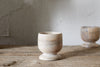 The Awa Wooden Egg Cup - Natural (Set of 2) by nkuku features small, lightweight wooden cups, ideal for your breakfast table and styled on a rustic surface with a textured off-white backdrop.