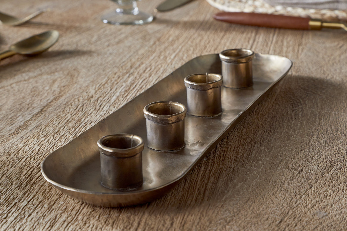 The nkuku Amita Long Candle Holder in antique brass features a rectangular design with rounded corners and four small cylindrical cups in a row, creating an elegant centrepiece for your dining table.
