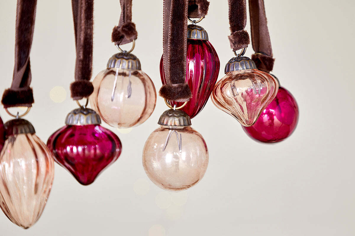 The Adisa Bauble Jar - Ruby & Copper (Set of 16) by nkuku features vintage-style mini baubles in varied shapes and finishes, hung from velvet ribbons for a charming festive accent against a soft, light backdrop.