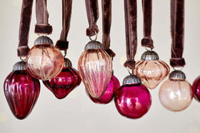 Close-up of nkuku’s Adisa Bauble Jar - Ruby & Copper (Set of 16): vintage-inspired glass ornaments in round and teardrop shapes, with ribbed textures in rich ruby and copper hues, hang from brown velvet ribbons for elegant festive décor.