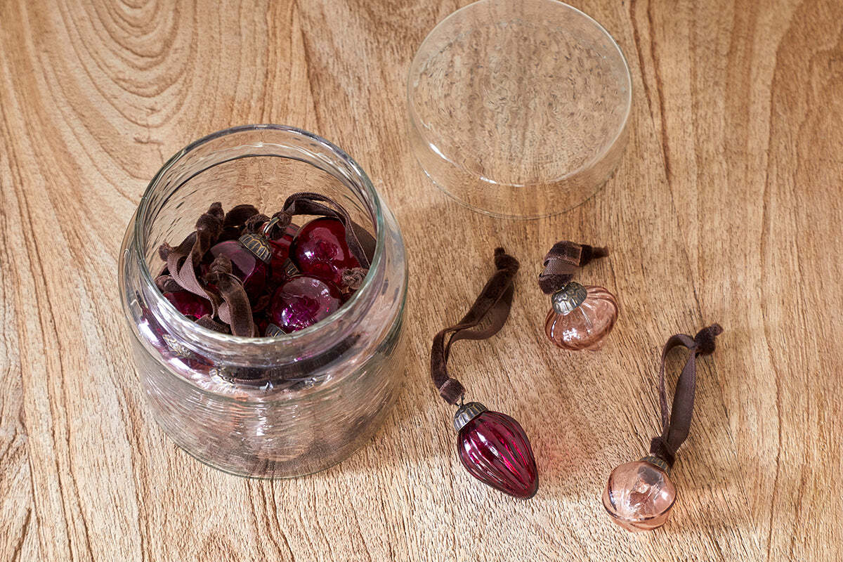 The nkuku Adisa Bauble Jar - Ruby & Copper (Set of 16) features mini glass baubles in festive shades, stored in a recycled glass jar. Some ornaments sit outside the open jar, making it perfect for adding holiday charm to any space.