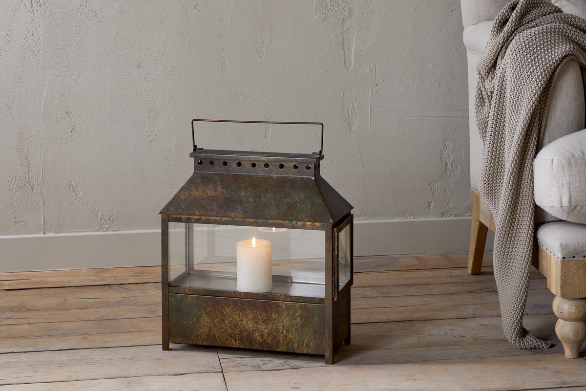 The Akoko Wide Lantern by nkuku, featuring a brown patina and glowing white pillar candle, adds rustic charm beside a beige upholstered chair with a knitted blanket against a textured light-toned wall.