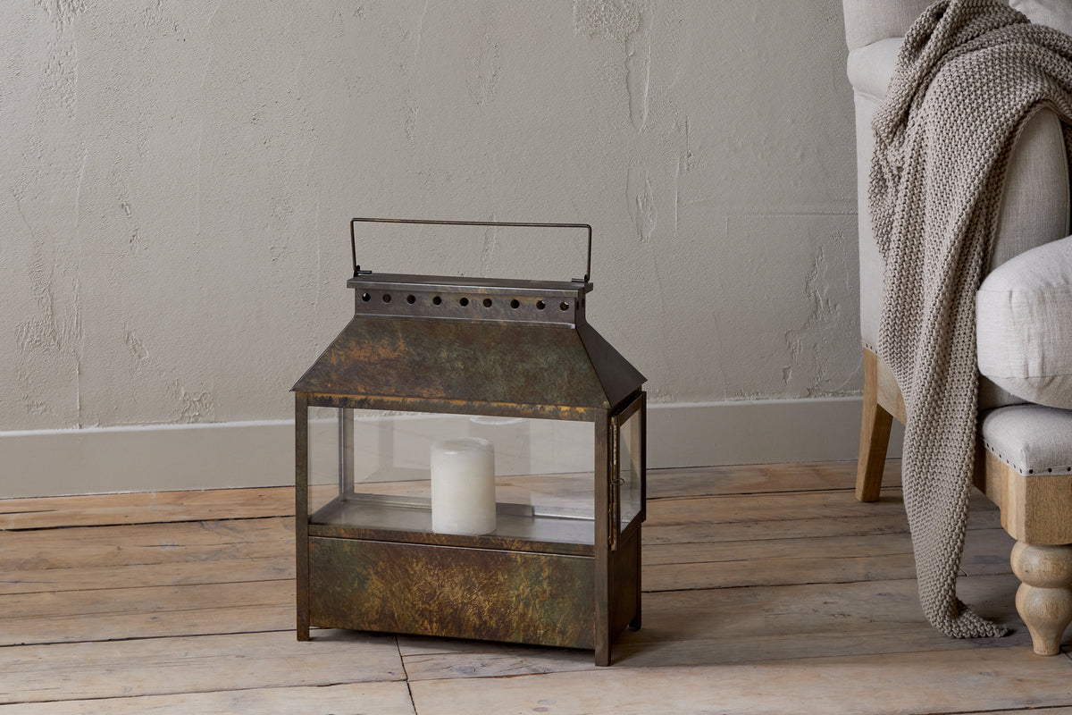 The Akoko Wide Lantern by nkuku, featuring a brown patina and glass panels, holds a white candle on a wooden floor next to a beige upholstered chair with a gray knitted blanket against a textured light gray wall.