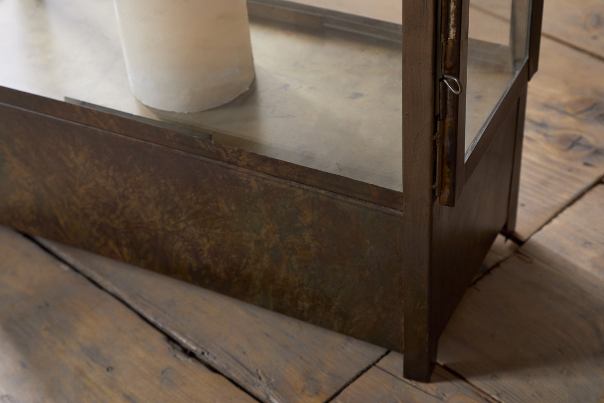 Close-up of the Akoko Wide Lantern by nkuku, featuring a brown patina finish on glass and metal, with a candle inside, placed on a rustic wooden floor.