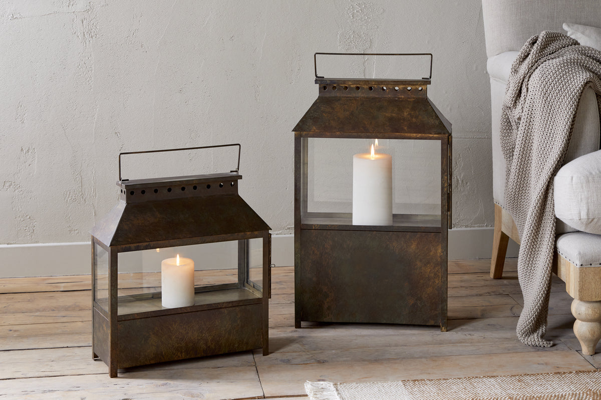 Two square Akoko Wide Lanterns by nkuku, finished in brown patina and each holding a lit white pillar candle, rest on a wooden floor. A beige armchair with wooden legs and a knitted throw sits against a textured neutral wall.