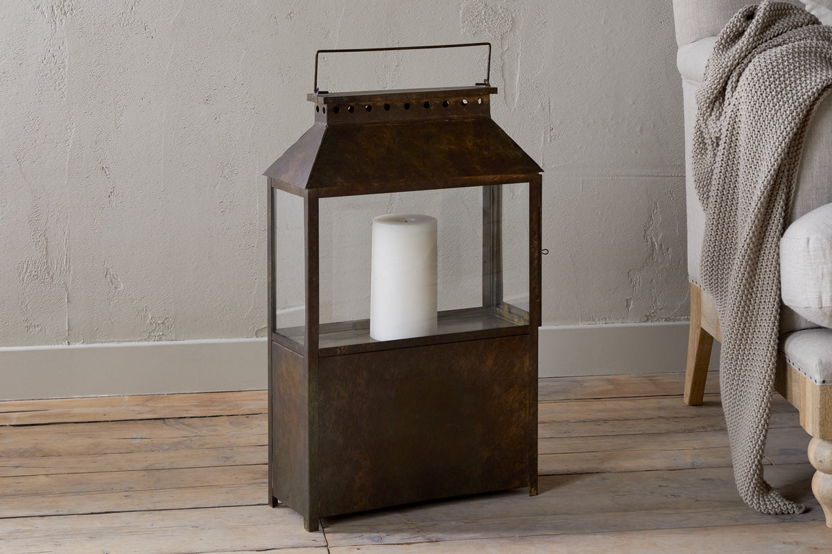 The nkuku Akoko Wide Lantern in Brown Patina with glass panels and a white pillar candle adds cozy charm beside a light-upholstered chair layered with a knit blanket.
