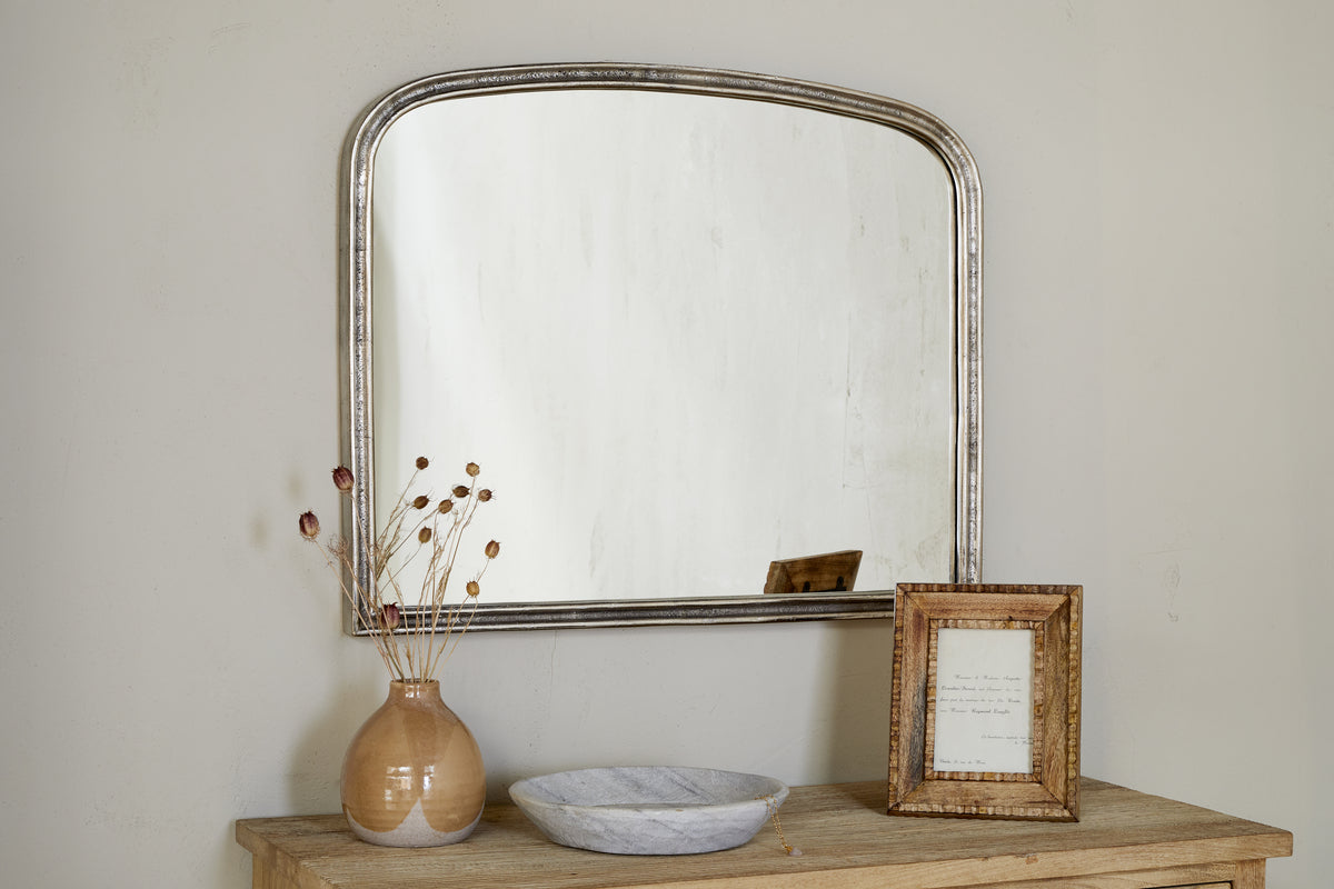 A wooden table with a beige vase of dried flowers, marble dish, and framed photo stands against the wall topped with nkukus Almora Wall Mirror in Antique Nickle, which features an arched design and elegant aluminium frame.