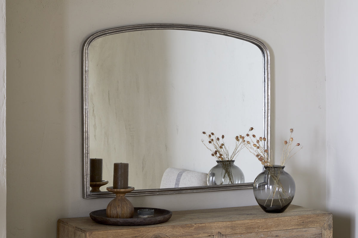 The nkuku Almora Wall Mirror in Antique Nickel, with its arched aluminium frame, hangs on a light wall above a rustic wooden table. A gray glass vase with dried flowers and a wooden tray with two candles decorate the table.