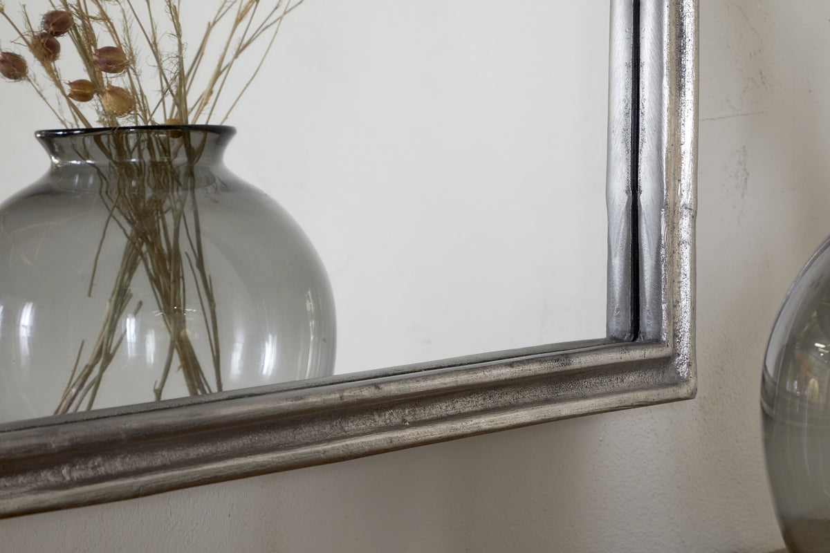 A close-up of dried plants in a gray glass vase, partially reflected in the nkuku Almora Wall Mirror (Antique Nickle) against a white wall.