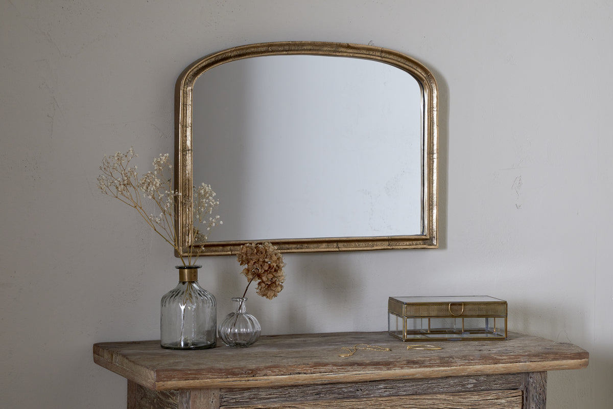 A rustic wooden table with two glass vases of dried flowers, a glass jewelry box, and the nkuku Almora Wall Mirror in Antique Brass (Extra Small) hanging on the light-colored wall above.